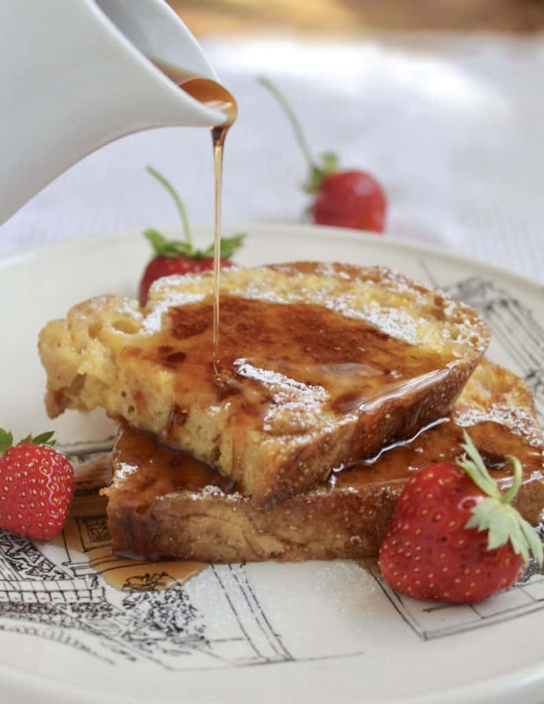 maple syrup being poured over slices of French toast on a plate with berries