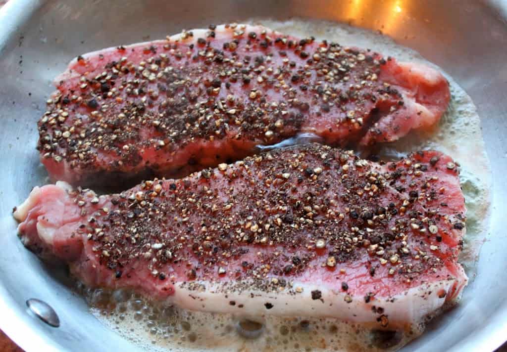 raw steaks just added to a pan with peppercorns