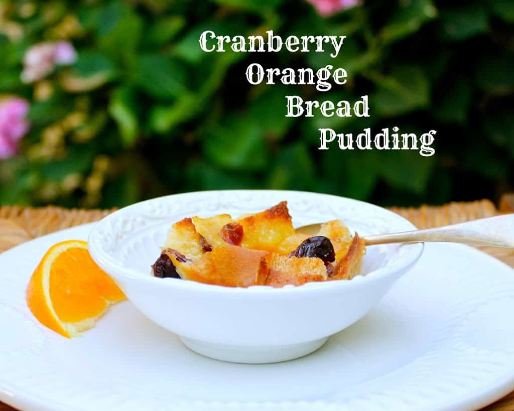 cranberry orange bread pudding in a white bowl with spoon and slice of orange and text 