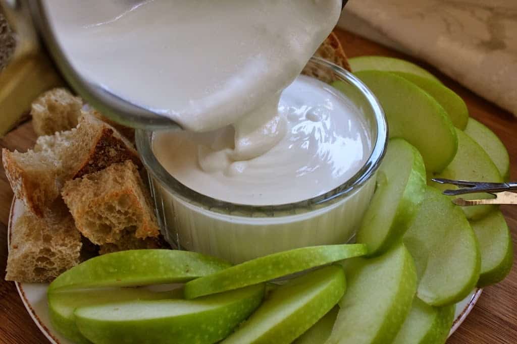 pouring Goat cheese fondue dip into a ramekin with bread and green apple slices on the plate